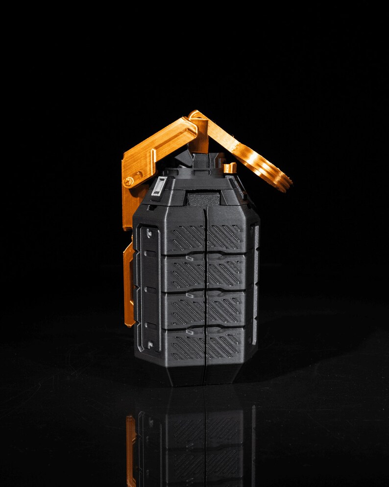 May include: A black and gold grenade with a textured surface and a gold lever. The grenade is on a black surface.