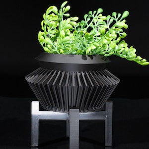 Pode incluir: Um vaso preto com um design canelado fica em cima de um suporte de metal preto. O vaso está cheio de plantas verdes de folha.