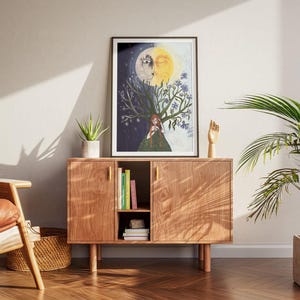 Puede incluir: Una l&aacute;mina de arte mural enmarcada que muestra un rostro dividido en sol y luna sobre un &aacute;rbol con una persona de pie debajo. El lado izquierdo es oscuro con estrellas, mientras que el lado derecho es claro con flores azules. El cuadro est&aacute; sobre un mueble.
