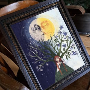 Puede incluir: Un cuadro enmarcado de 30x40 cm que muestra un rostro dividido en sol y luna sobre una persona de largo cabello rojo. La persona est&aacute; frente a un &aacute;rbol con flores azules, sobre un fondo de cielo estrellado azul oscuro y un fondo amarillo claro.