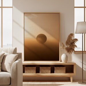 May include: A large framed artwork depicts a gradient of warm brown tones with a central circular shape, resembling a sunset or eclipse. The artwork is displayed above a wooden console with woven storage baskets, complemented by a vase of dried pampas grass and a floor lamp.