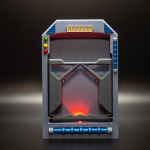 May include: A futuristic-looking, rectangular device with a gray frame, blue accents, and a red light emanating from the center. It features a clear panel with geometric shapes and a series of colored lights.
