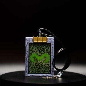 May include: A rectangular pendant with a black cord. The pendant has a gray frame with a gold-colored top and a green design on a black background. The design is a squiggly green shape within a circuit board pattern.