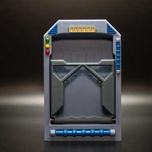 May include: A gray and blue rectangular device with a geometric design. It features a central opening with a stylized emblem, accented by blue and gold details. The device has a series of colored lights on the side.