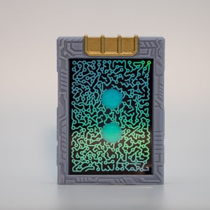 May include: A rectangular, gray device with a gold accent at the top. The screen displays a circuit board design with two teal orbs. The device has a futuristic aesthetic.