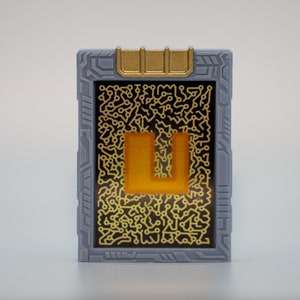 May include: A rectangular, gray device with a gold-colored top and a black screen. The screen features a gold circuit board design and a gold-colored letter "U". The device has a futuristic, tech-inspired aesthetic.