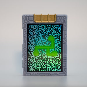 May include: A rectangular device with a gray frame and a gold-colored top. The screen displays a circuit board pattern in shades of blue and green, with a bright green, stylized shape in the center.