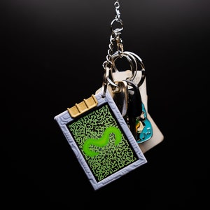 May include: A keychain with a rectangular charm featuring a green squiggly design on a black background, framed in gray. The charm has a gold-colored top and is attached to a keyring with other keys and charms. The background is black.
