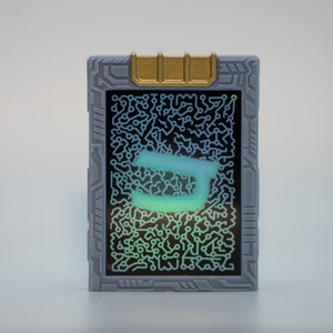 May include: A rectangular, gray device with a gold accent at the top. The screen displays a glowing, teal-colored letter "J" against a backdrop of intricate circuit patterns. The device has a futuristic, tech-inspired design.