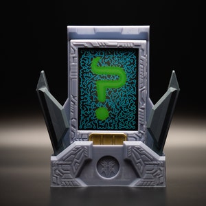 May include: A gray, futuristic-looking device with a screen displaying a green question mark. The device has angular, geometric designs and a gold-colored accent at the base of the screen. The background is dark.