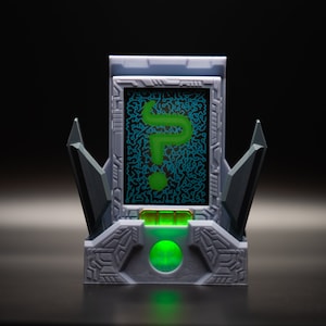 May include: A futuristic-looking device with a gray and black color scheme. The device features a screen displaying a green question mark on a circuit board background. The base has a glowing green orb. Two angular, dark gray structures flank the screen.