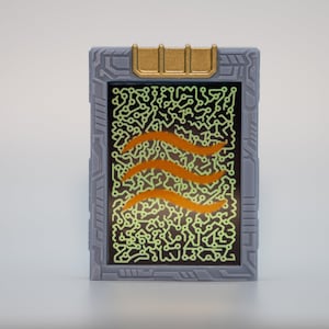 May include: A gray rectangular device with a gold top and a screen displaying a green circuit pattern and three orange wavy lines. The device has a futuristic design.