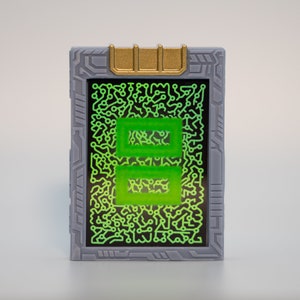 May include: A rectangular device with a gray frame and a gold-colored top. The screen displays a green circuit board design with two green rectangles. The device has a futuristic, tech-inspired aesthetic.