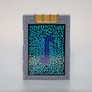 May include: A rectangular device with a gray frame and a gold-colored top. The screen displays a blue circuit board design with a blue "J" shape in the center. The device has a futuristic aesthetic.
