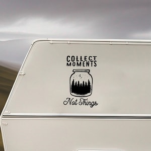May include: A white travel trailer featuring a black graphic decal on its side. The decal displays a jar silhouette filled with a forest scene, accompanied by the text "COLLECT MOMENTS" above and "Not Things" below in a stylized black font.