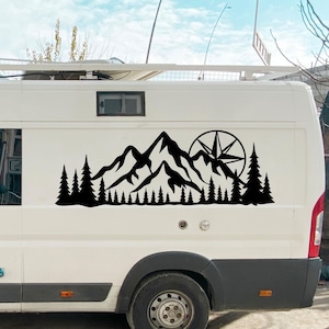 Op de afbeelding: Zwarte berg- en bossticker op een witte bestelwagen. Het ontwerp toont een bergketen, dennenbomen en een kompasroos. Ideaal voor outdoorliefhebbers.