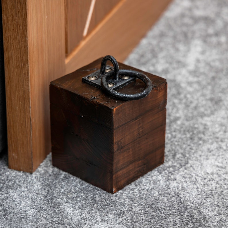 Dark wooden doorstop created from four layers of wood, with a black metal hoop on top attached with brass screws stands in front of an oak door on grey carpet.