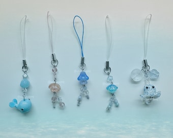 May include: Five beaded charms with ocean-themed designs. The charms include a blue whale, jellyfish, and octopus, with clear and pastel beads. Each charm has a silver-tone metal loop and a clear lanyard.
