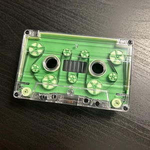 Op de afbeelding: Een doorzichtige plastic cassetteband met een felgroen binnenframe. Het ontwerp bevat twee ronde spoelen, meerdere kleine geleidingswieltjes en een centraal rechthoekig venster, alles ingesloten in een transparante behuizing met vier schroeven.