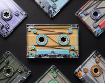 May include: A collection of six transparent cassette tapes arranged in a circular pattern. The central tape features a tan interior with bright turquoise spools and a clear outer shell. Other tapes display purple, blue, red, olive, and grey accents.