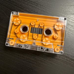May include: A rectangular audio cassette tape featuring a clear plastic outer shell and a vibrant orange inner casing. The design displays visible internal spools, guide rollers, and magnetic tape winding through the mechanism with two circular hubs.