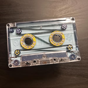 May include: A clear cassette tape with a light blue interior and yellow and purple accents. The tape has a ghost-like graphic on the front. The tape is on a dark brown surface.