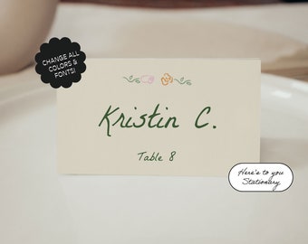 Pode incluir: Um cartão de lugar branco com texto verde que diz "Kristin C." e "Mesa 8". O cartão tem um design floral com detalhes rosa, laranja e verde. O texto "Here's to you Stationery" está escrito em verde sobre um oval branco.
