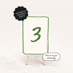 May include: Green and white table number card with the number 3 written in green. The card is on a small wooden easel. The text "Here's to you stationery" is written in black below the card.