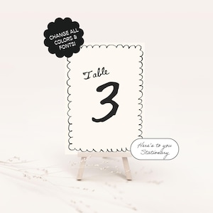 May include: Black and white table number card with the number 3 and the text "Table" on a white background. The card is on a small wooden easel. The text "Here's to you Stationary" is on a black and white sticker.