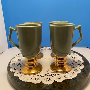 May include: Four olive green coffee mugs with gold-colored pedestal bases and handles. The mugs are arranged on a marble-like surface with a white lace doily. The background is a solid blue.