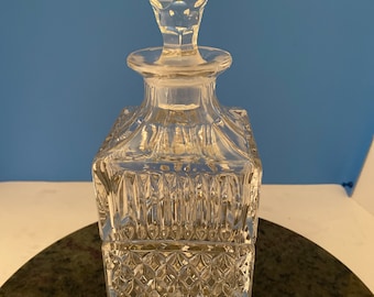 May include: A clear glass decanter with a square body and a faceted design. The decanter has a clear glass stopper with a faceted design.