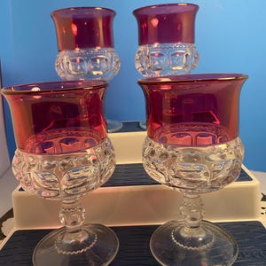 May include: Four vintage wine glasses with a clear, textured base and a vibrant, ruby-red bowl. The rim of each glass is accented with a thin gold band. The glasses are arranged on a white and blue surface, with a blue background.