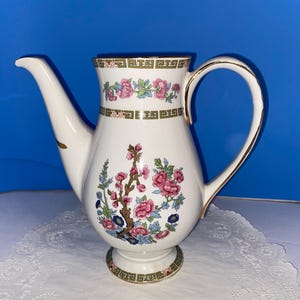 May include: White ceramic teapot with a floral design featuring pink flowers, blue accents, and green leaves. The teapot has a curved spout, a gold-trimmed handle, and a decorative band around the top. The base also has a decorative band.