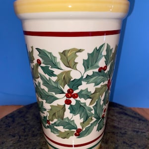 May include: A tall, ceramic vase with a cream-colored base and a yellow rim. The vase is decorated with green holly leaves and red berries. A red stripe encircles the vase near the top and bottom.
