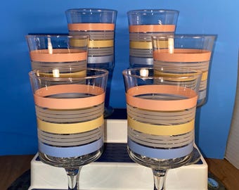 May include: Six vintage wine glasses with colorful horizontal stripes in peach, yellow, gray, and blue. The glasses are clear with a stem and a round base. The glasses are arranged on a white and blue tiered display.