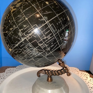 May include: A black and gold globe with a detailed world map, mounted on a gray marble base. The globe has a copper-colored meridian and a small copper sphere at the top. The globe is approximately 10 inches tall.