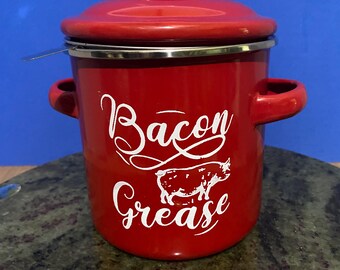 May include: A bright red cylindrical metal grease container with a matching lid and two side handles. The front features white cursive text that reads Bacon Grease, accompanied by a white graphic illustration of a pig silhouette.