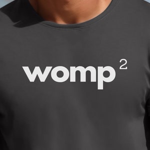 Puede incluir: Camiseta gris oscuro con la palabra "womp²" impresa en blanco. La camiseta tiene mangas cortas y cuello redondo. El fondo muestra una vista borrosa del océano y palmeras.