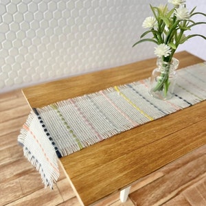May include: A white woven table runner with pink, yellow, and blue stripes on a wooden table with a vase of white flowers.