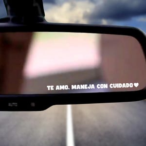 May include: A car's rearview mirror with the text "TE AMO, MANEJA CON CUIDADO" in white, along with a heart symbol. The mirror reflects a road and a cloudy sky.