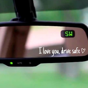 May include: A car's rearview mirror with the text "I love you, drive safe" in white script. The mirror has a green digital display and a green light. The background is a blurred view of greenery.