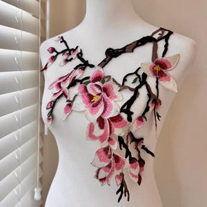 May include: Embroidered floral applique featuring pink and white blossoms with yellow centers, and pink buds, attached to brown branches. The applique is displayed on a white mannequin, with a window and blinds in the background.