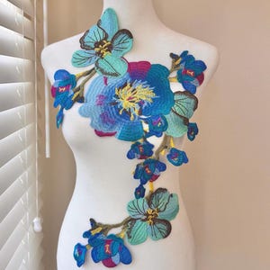 Op de afbeelding: Geborduurde bloemenapplicatie in tinten blauw, turquoise en magenta, gepresenteerd op een witte mannequin. Het ontwerp toont een grote centrale bloem met kleinere bloesems die naar beneden vallen. De applicatie is waarschijnlijk voor kleding of knutselprojecten.