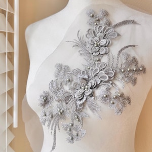 May include: A silver floral applique with sequins and faux pearls. The applique features a detailed design of flowers and leaves, with small pearl accents. The applique is attached to a sheer, white fabric backing, and is displayed on a mannequin.
