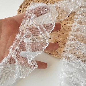 May include: Close-up of white lace trim with a scalloped edge and small, evenly spaced white polka dots. The delicate fabric is held against a natural woven background, showcasing the trim's texture and design. Suitable for crafting and sewing projects.