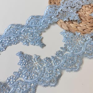 May include: A light blue lace trim featuring intricate floral embroidery and scattered clear sequins. The delicate fabric has scalloped edges and a detailed pattern of interconnected blossoms, providing a decorative border for sewing projects.