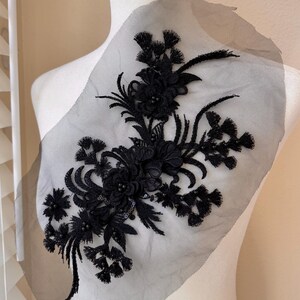May include: A black floral lace applique featuring intricate embroidery, small beads, and sequins on a sheer black mesh backing. The design displays a central cluster of flowers with radiating stems, leaves, and fringe-like accents in black thread.
