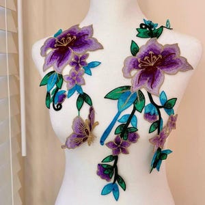 Op de afbeelding: Paarse en blauwe geborduurde bloemenapplicatie met een vlindermotief. De applicatie is perfect om kleding, tassen of woonaccessoires een vleugje elegantie te geven.