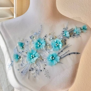 May include: A delicate floral applique featuring light blue flowers and silver embellishments. The applique is displayed on a white dress form.