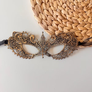May include: Ornate black and gold masquerade mask with intricate filigree detailing. The mask features a central crown design and is attached to black ribbons. The mask is set against a neutral background with a woven texture.